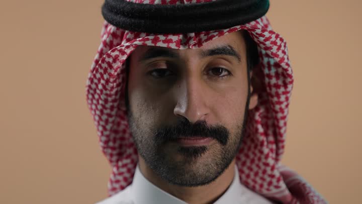 الهدوء والسكون وعدم الإزعاج، بورتريه مقرب لشاب عربي خليجي سعودي يرتدي الثوب الابيض والشماغ يضع أصبع السبابة على فمه، التوقف عن الكلام وخفض الصوت، طلب السكوت والصمت، حركة أصبع وإشارات، خلفية بنية