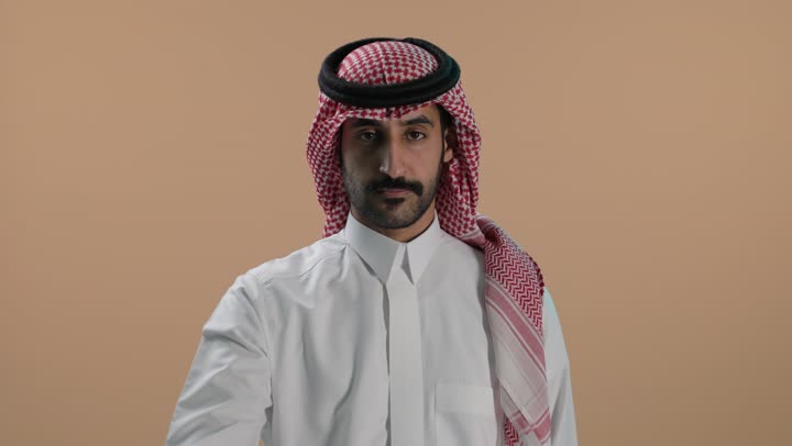 مفهوم الرفض والامتناع والانزعاج, بورتريه لشاب عربي خليجي سعودي يرتدي الثوب الابيض والشماغ يمد يده نحو الأمام مع رفع الكف بإيماءات النهي عن فعل امر ما, طلب التوقف وعدم الاقتراب, تعابير الحزم والجدية, خلفية بنية