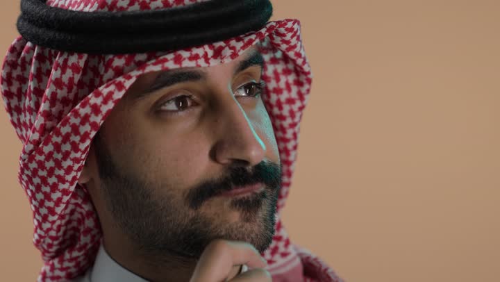 التأمل وشرود الذهن، التفكير في شيء هام، , بورتريه مقرب لشاب عربي خليجي سعودي يرتدي الثوب الابيض والشماغ يضع يده على ذقنه وينظر الى شيء، خلفية بنية