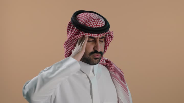 الشعور بالإرهاق وفقدان التركيز, النسيان ومحاولة تذكر شيء ما, الاحساس بالشقيقية والصداع النصفي, بورتريه لشاب عربي خليجي سعودي يرتدي الثوب الابيض والشماغ يضع يديه على رأسه بإيماءات وجع في الدماغ والرأس الشديد, خلفية بنية