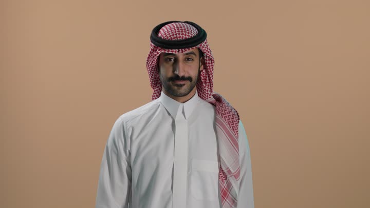 مفهوم العناية والتجميل والتبيض، بورتريه لشاب عربي خليجي سعودي يرتدي الثوب الابيض والشماغ ينظر إلى الكاميرا يشير بيديه إلى أسنانه, تعابير السرور باستخدام قشور فينير من الخزف، أهمية متابعة طبيب الاسنان، خلفية بنية