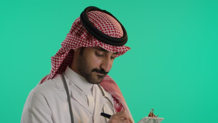 التفكير و محاولة تذكر شيء ، العمل في المراكز والمستشفيات، الطب والرعاية الصحية، تدوين الملاحظات والبيانات، بورتريه لطبيب عربي خليجي سعودي يرتدي الشماغ و المعطف الطبي الأبيض يضع سماعة الفحص الطبية يقوم بالكتابة، خلفية خضراء ، كروما