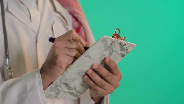 Working in centers and hospitals, medicine and healthcare, taking notes and data, a close-up portrait of the hand of an Arab Gulf Saudi man wearing a white medical coat and using a stethoscope, writing something, green background, chroma.