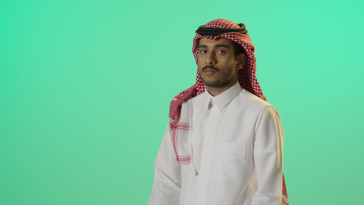 The concept of caring about external appearance, masculinity, and elegance, gestures of self-confidence, a portrait of a young Arab Gulf Saudi man wearing a white thobe and a shemagh standing upright with crossed arms, looking at the camera with expressions of seriousness and determination, green background, chroma.