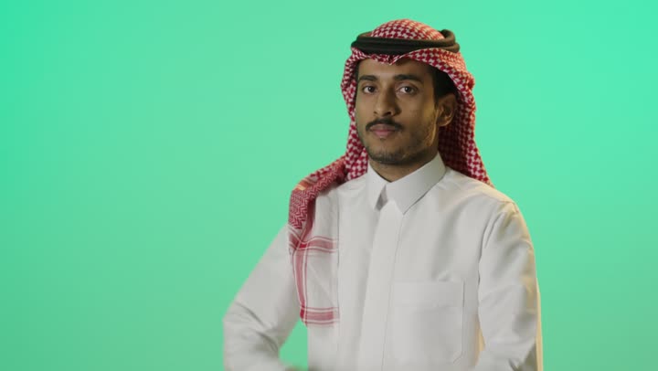 ايماءات الثقة بالنفس، مفهوم الاهتمام بالمظهر الخارجي و الرجولة والأناقه، بورترية لشاب عربي خليجي سعودي يرتدي الثوب الابيض و الشماغ يقف باستقامة بايدي متقاطعه, النظر إلى الكاميرا بتعابيرالجدية والحزم، خلفية خضراء ، كروما