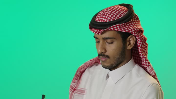تدوين وتسجيل الملاحظات والمعلومات على الورق، إدارة الأعمال وتنظيمها, بورترية لشاب عربي خليجي سعودي يرتدي الثوب الابيض و الشماغ يقوم بكتابة المهام والأهداف اليومية، خلفية خضراء ، كروما