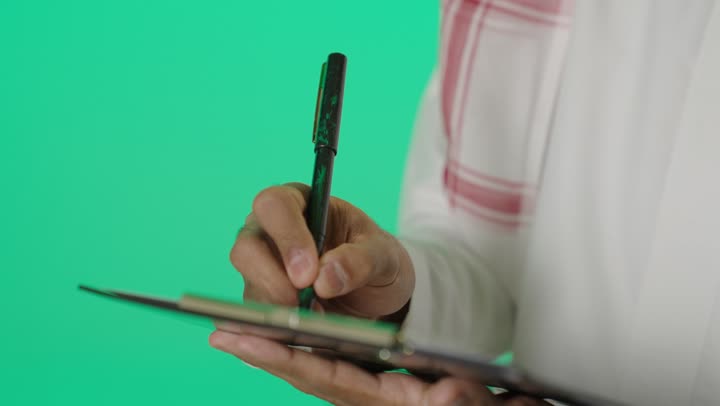 Business management and organization, writing and recording notes and information on paper, a close-up portrait of a young Arab Gulf Saudi man wearing a white thobe and a shemagh writing daily tasks and goals, green background, chroma.