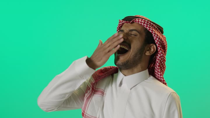 Laziness and lethargy and the desire for rest, staying up late, a close portrait of a young Arab Gulf Saudi man wearing a white thobe and a shemagh, placing his hands on his mouth as he yawns, green background, chroma.