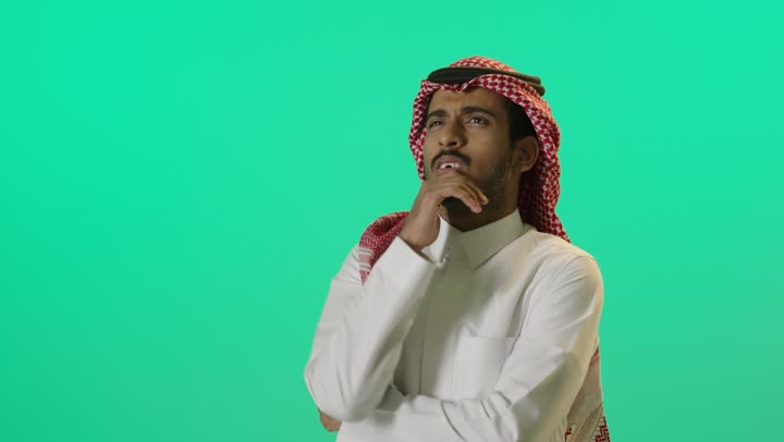 Focus, distraction, and contemplation, to reflect and plan for the future, finding a solution to a problem, a close-up portrait of a young Arab Gulf Saudi man wearing a white thobe and a shemagh, resting his hand on his chin, looking up with gestures of thought and concentration, green background, chroma.