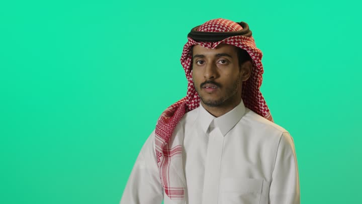 Greeting and peace, elegance, handsomeness, and masculinity, the concept of appreciation and respect, a close portrait of a young Arab Gulf Saudi man wearing a white thobe and a shemagh raising his right hand to perform a military salute, green background, chroma.