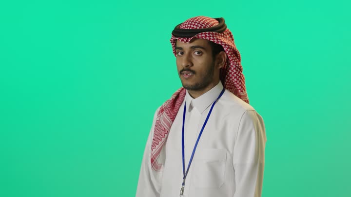 ايماءات الثقة بالنفس، مفهوم الاهتمام بالمظهر الخارجي و الرجولة والأناقه، بورترية لشاب عربي خليجي سعودي يرتدي الثوب الابيض و الشماغ و بطاقة تعريفية بشريط ازرق يقف باستقامة بايدي متقاطعه, النظر إلى الكاميرا بتعابيرالسعادة و السرور، خلفية خضراء، كروما
