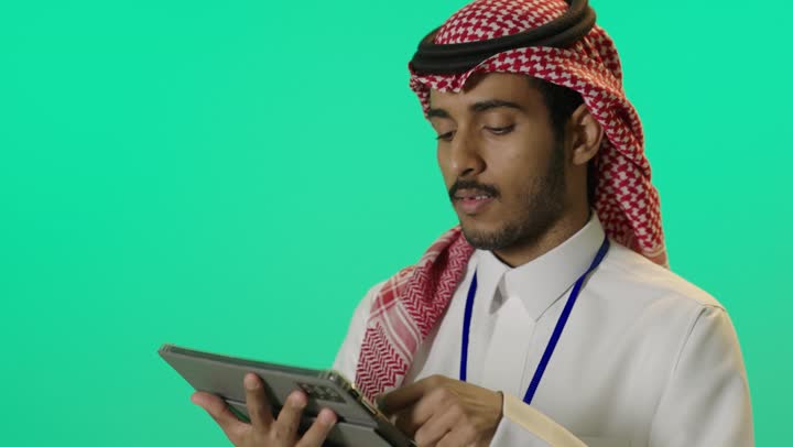 Tablet programs and applications, chatting with family and friends, using technology and modern techniques, a portrait of a young Arab Gulf Saudi man wearing a white thobe and a shemagh with an identification card with a blue strip, writing on a tablet device, browsing social media programs, green background, chroma.