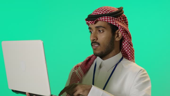 تصفح مواقع التواصل الإجتماعي ،ادارة شؤون الاعمال وتنظيمها، استخدام الأجهزة التقنية الحديثة والمتطورة، العمل عن بعد، بورترية لشاب عربي خليجي سعودي يرتدي الثوب الابيض و الشماغ و بطاقة تعريفية بشريط ازرق يستخدم الحاسوب المحمول، خلفية خضراء، كروما