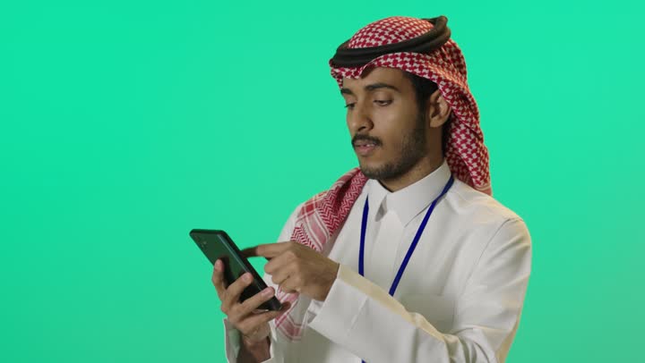 استخدام وسائل التواصل الرقمية، التحدث والدردشة مع الأصدقاء عبر الجوال، بورترية لشاب عربي خليجي سعودي يرتدي الثوب الابيض و الشماغ و بطاقة تعريفية بشريط ازرق يستخدم الهاتف المحمول، خلفية خضراء ، كروما