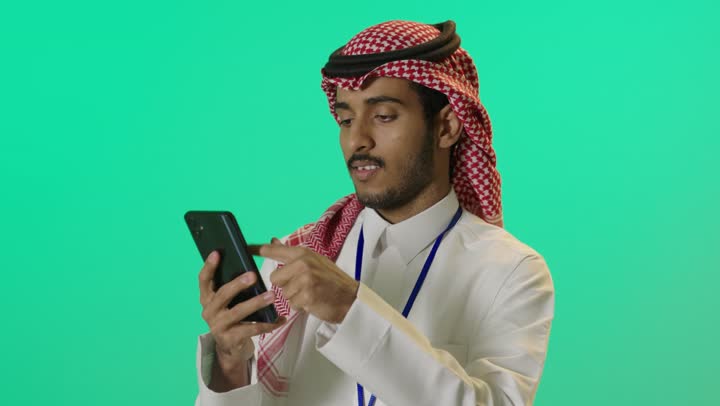 Following social networks, talking and chatting with friends via mobile, a portrait of a young Arab Gulf Saudi man wearing a white thobe and a shemagh, with an identification card with a blue stripe using a mobile phone, green background, chroma.