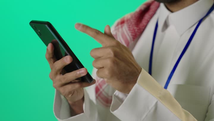 التحدث والدردشة مع الأصدقاء عبر الجوال، تصفح وسائل التواصل الاجتماعي، بورترية مقرب ليد شاب عربي خليجي سعودي يرتدي الثوب الابيض و الشماغ و بطاقة تعريفية بشريط ازرق يستخدم الهاتف المحمول، خلفية خضراء ، كروما