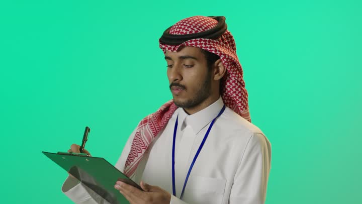 تدوين المعلومات و الملاحظات المهمة، التفكير فى شيء ما، بورتريه لشاب عربي سعودي خليجي يرتدي الثوب التقليدي و الشماغ وبطاقة هوية تعريفية خاصة بالعمل يقوم بكتابة شيء ما, خلفية خضراء، كروما