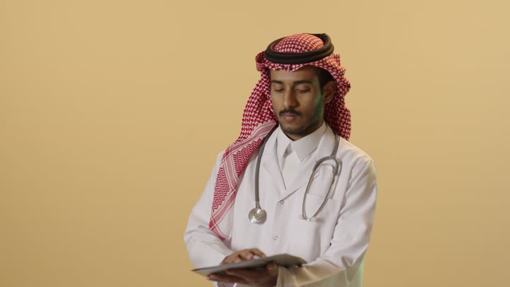 قراءة نتائج الفحوصات بالسجل الطبي,بورتريه لطبيب عربي خليجي سعودي يرتدي الزي التقليدي والشماغ والمعطف الطبي يقوم  بإعداد التقارير الطبية, تشخيص الحالات المرضية, خلفية بيج