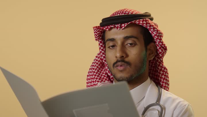تشخيص الحالات المرضية، قراءة نتائج الفحوصات بالسجل الطبي,بورتريه مقرب لطبيب عربي خليجي سعودي يرتدي الزي التقليدي والشماغ والمعطف الطبي يقوم  بإعداد التقارير الطبية, خلفية بيج