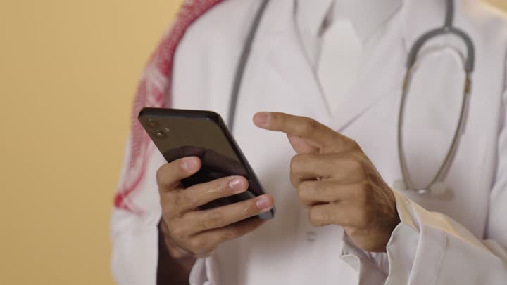 التطبيقات الرقمية للخدمات الصحية, تقديم الخدمات والرعاية الصحية عبر الإنترنت، بورتريه مقرب ليد طبيب عربي خليجي سعودي يرتدي الزي التقليدي والشماغ والمعطف الطبي يقوم باستخدام الهاتف المحمول، استخدام الأجهزة التقنية الحديثة ودمج التكنولوجيا مع الطب، خلفية بيج