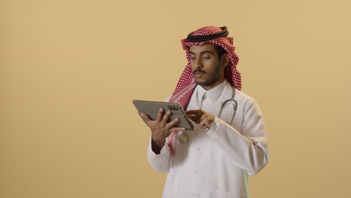 التطور التقني في مختلف المجالات, استخدام التكنولوجيا الحديثة في العمل عن بعد، إنجاز الأعمال بسرعة وسهولة، بورتريه مقرب لطبيب عربي خليجي سعودي يرتدي الزي التقليدي والشماغ والمعطف الطبي يحمل بيده جهاز التابليت, خلفيه بيج