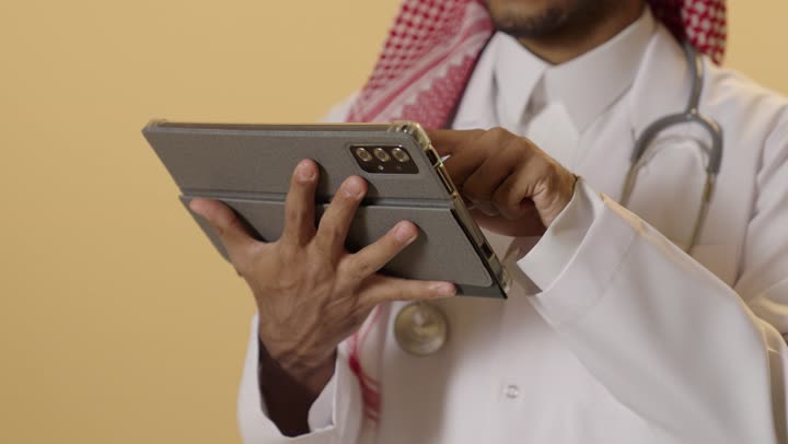 The use of modern technology in remote work, technological advancement in various fields, accomplishing tasks quickly and easily, a close-up portrait of a Saudi Arabian Gulf Arab doctor wearing traditional attire, a ghutrah, and a medical coat holding a tablet in his hand, with a beige background.