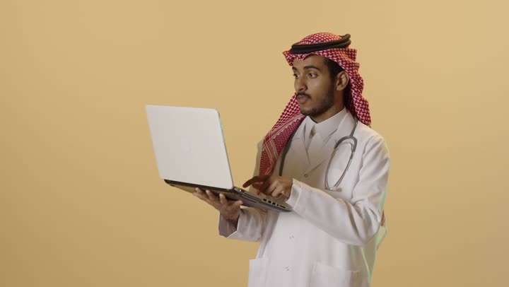 إدارة ملفات المرضى إلكترونيًا على اللابتوب, دمج التقنية الحديثة في مجال العمل الطبي, بورتريه مقرب لطبيب عربي خليجي سعودي يرتدي الزي التقليدي والشماغ والمعطف الطبي يعمل على الحاسب المحمول, تقديم الخدمات العلاجية في المراكز الطبية والمستشفيات , خلفية بيج