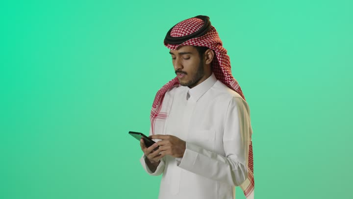 التواصل عبر الإنترنت، بورترية لشاب عربي خليجي سعودي يرتدي الثوب الابيض و الشماغ يستخدم الهاتف المحمول بايماءات الاندماج و التركيز، تصفح التطبيقات الذكية، استخدام التقنيات الحديثة ، خلفية خضراء، كروما