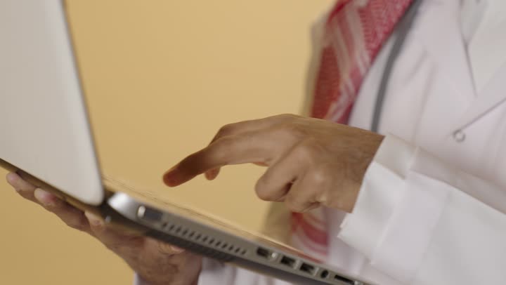 دمج التقنية الحديثة في مجال العمل الطبي، إدارة ملفات المرضى إلكترونيًا على اللابتوب, بورتريه مقرب ليد طبيب عربي خليجي سعودي يرتدي الزي التقليدي والشماغ والمعطف الطبي يعمل على الحاسب المحمول, تقديم الخدمات العلاجية في المراكز الطبية والمستشفيات , خلفية بيج
