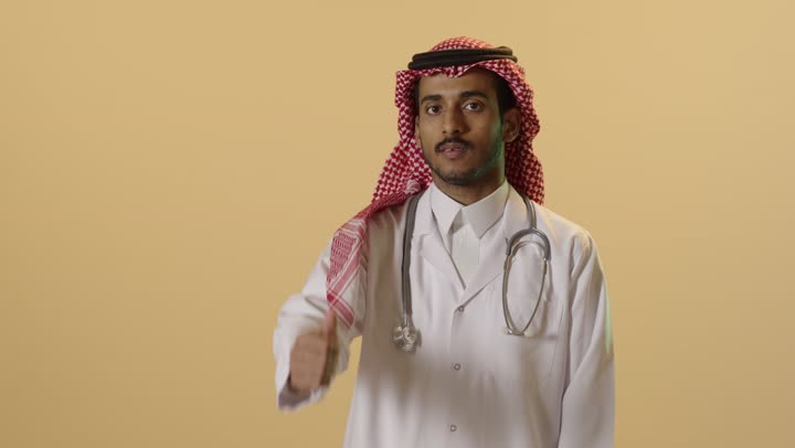 Satisfaction with medical services and healthcare, a close-up portrait of an Arab Gulf Saudi doctor wearing traditional attire, a ghutrah, and a medical coat, raising his thumb with gestures of admiration and satisfaction for achieving a certain matter, with a brown background.