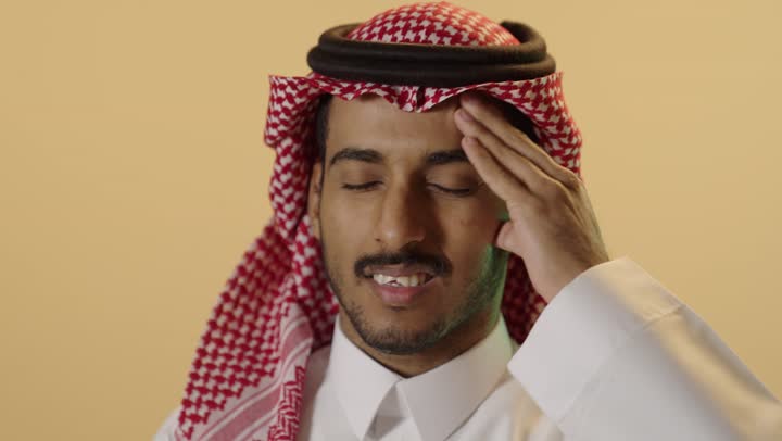 آلام شديدة في الرأس، ايماءات وجه ويدين تدلان على الشعور بالألم، بورترية مقرب لشاب عربي خليجي سعودي يرتدي الثوب الابيض و الشماغ يشير بيديه الى رأسه بأيماءات المرض والاحساس بالصداع والحاجه الى العلاج والراحه، خلفيه بيج