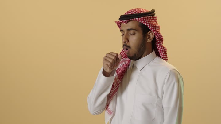 إيماءات الإرهاق والتعب الشديد،الاصابة بفايروس كورونا، الإصابة بالأنفلونزا ونزلات البرد، الكحة والتهاب الحلق والحاجة للعلاج، بورترية لشاب عربي خليجي سعودي يرتدي الثوب الابيض و الشماغ يضع يده أمام فمه ويقوم بالسعال, خلفية بيج