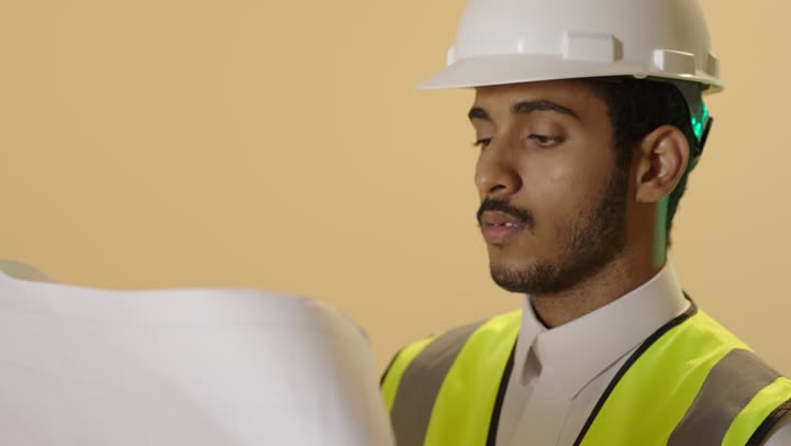 مفهوم ادارة المشاريع وتدقيقها، العمل في القطاع الهندسي، بورتريه مقرب لمهندس عربي خليجي سعودي يرتدي الثوب الابيض والخوذة وسترة الحمايةيقوم بمتابعة الاعمال ومخططات المشروع، خلفية بيج