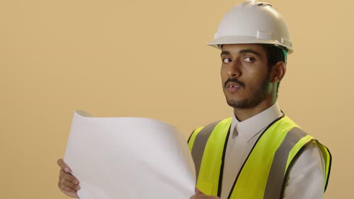 العمل في القطاع الهندسي، مفهوم ادارة المشاريع وتدقيقها، بورتريه مقرب لمهندس عربي خليجي سعودي يرتدي الثوب الابيض والخوذة وسترة الحمايةيقوم بمتابعة الاعمال ومخططات المشروع، خلفية بيج