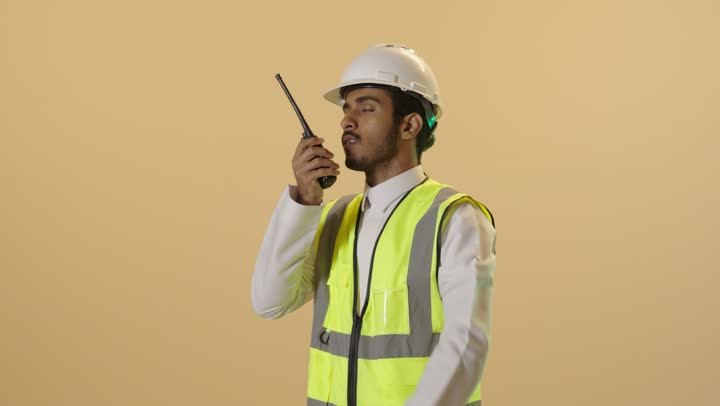 المتابعة والإشراف على خطط العمل، بورتريه مقرب لمهندس عربي خليجي سعودي يرتدي الثوب الابيض والخوذة وسترة الحماية يقوم باستخدام اللاسلكي، مهن و وظائف هندسية، ادارة المشاريع المعمارية الهندسية، خلفية بنية