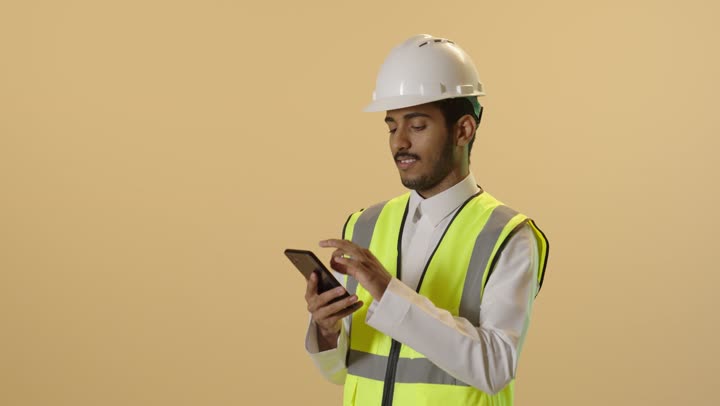 Integrating modern technology with engineering, professions and jobs for youth, working in the engineering sector, a close-up portrait of a Saudi Gulf Arab engineer wearing a white thobe, helmet, and safety vest, monitoring the progress of work remotely via a mobile phone, beige background.