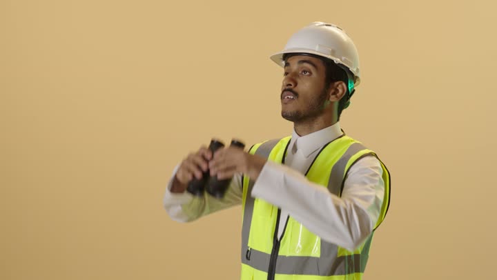 رؤية الأشياء البعيدة، بورتريه مقرب لمهندس عربي خليجي سعودي يرتدي الثوب الابيض  والخوذة وسترة الحماية ينظر من خلال المنظار ثنائي العينين إلى شيء ما ، متابعة سير المشاريع الهندسية الإعمارية، المهن والوظائف الشبابية، خلفية بيج