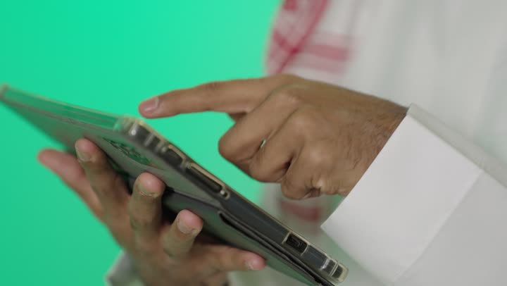 Following the latest news from social media sites online, a close-up portrait of the hand of a young Arab Gulf Saudi man with a dark complexion wearing a white thobe tapping on a tablet device, communicating and chatting with relatives and friends, remote electronic marketing, green background, chroma.