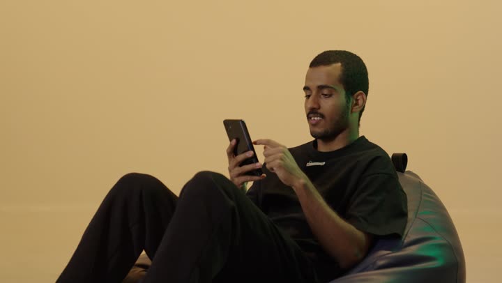 تصفح مواقع التواصل الاجتماعي عبر الجوال، المتعة والتسلية باستخدام التقنية الحديثة، إنجاز مهام العمل عن بعد، بورتريه لشاب عربي خليجي سعودي اسمر يرتدي الزي الكاجوال يجلس على كرسي بين باج  يقوم باستخدام الهاتف المحمول، خلفية بنية