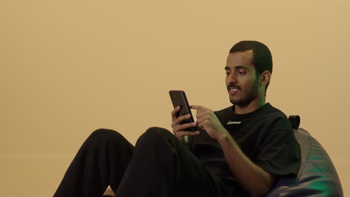 المتعة والتسلية باستخدام التقنية الحديثة، تصفح مواقع التواصل الاجتماعي عبر الجوال، إنجاز مهام العمل عن بعد، بورتريه لشاب عربي خليجي سعودي اسمر يرتدي الزي الكاجوال يجلس على كرسي بين باج  يقوم باستخدام الهاتف المحمول، خلفية بنية