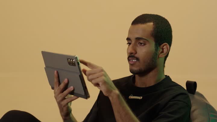 الدردشة والتحدث مع الاصدقاء عبر الانترنت، تصفح تطبيقات جهاز التابلت، بورتريه مقرب لشاب عربي خليجي سعودي اسمر يرتدي الزي الكاجوال يجلس على كرسي بين باج يقوم بمتابعة اخبار وسائل التواصل الاجتماعي باستخدام التابلت، خلفية بنية