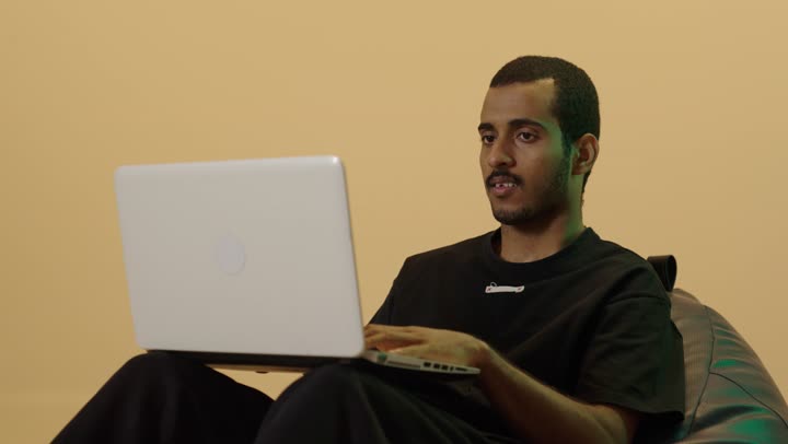 Browsing the internet and social media, completing work and tasks remotely via a laptop, a portrait of a young Arab Gulf Saudi man with a dark complexion wearing casual attire sitting on a chair between bags using a laptop, brown background.