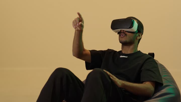 The enjoyment and entertainment of watching through virtual reality glasses, immersion and simulation of the imaginary world, a portrait of a young Arab Gulf Saudi man with a tan wearing casual attire sitting on a chair between bags wearing 3D glasses (VR), technological and technical development, pointing to something, brown background.