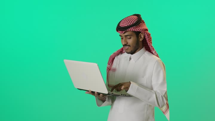 صناعة المحتويات النصية، النقر على لوحة المفاتيح، مفهوم ادخال البيانات، بورترية مقرب ليد شاب عربي خليجي سعودي يرتدي الثوب الابيض و الشماغ يقوم بالكتابة على جهازاللابتوب، دمج التكنولوجيا الحديثة بالحياة العملية، خلفية خضراء، كروما