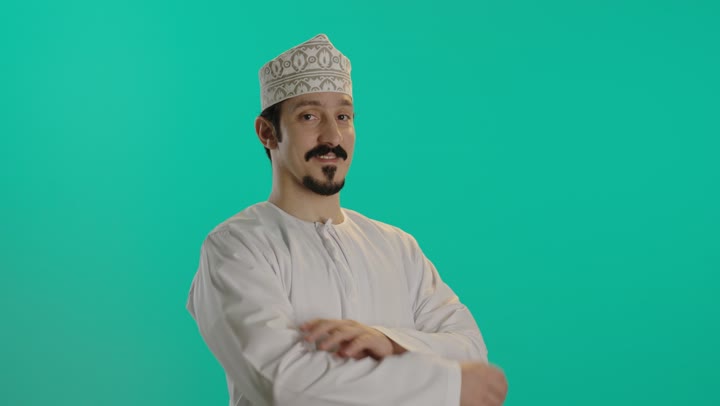 رجل عربي خليجي عماني يرتدي كمة مطرزة و دشداشة بيضاء يقف امام الكاميرا يعقد ذراعه في ثقة  , تعابير السعادة والابتسامة , الوقوف بعزة و شموخ