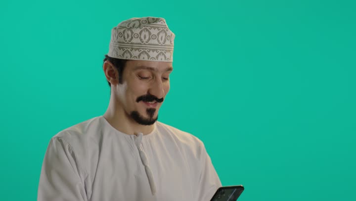 Spending enjoyable time with fun applications during leisure time, an Arab Gulf Omani man wearing embroidered cuffs and a white dishdasha holds a mobile phone, with expressions of happiness and joy while using various applications.
