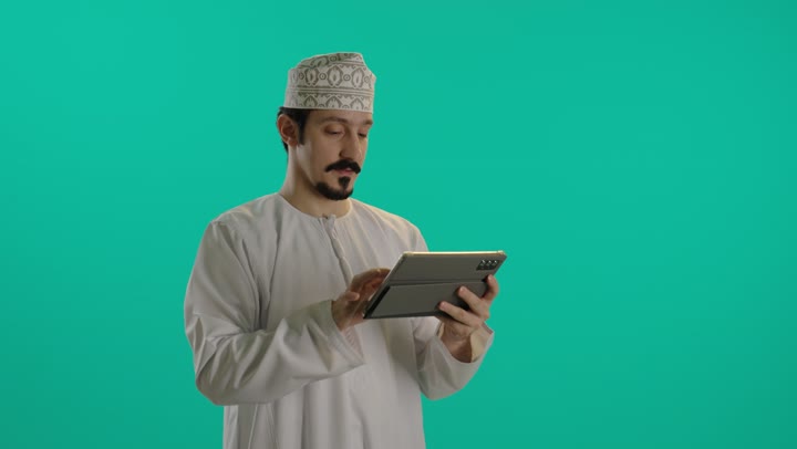 تعابير الجدية اثناء انجاز الاعمال المطلوبة بشكل مثالي , رجل عربي خليجي عماني يرتدي الدشداشة البيضاء و الكمة المطرزة يحمل الجهاز اللوحي الخفيف المتطور , تعابير السعادة و الابتسامة الواضحة امام الكاميرا