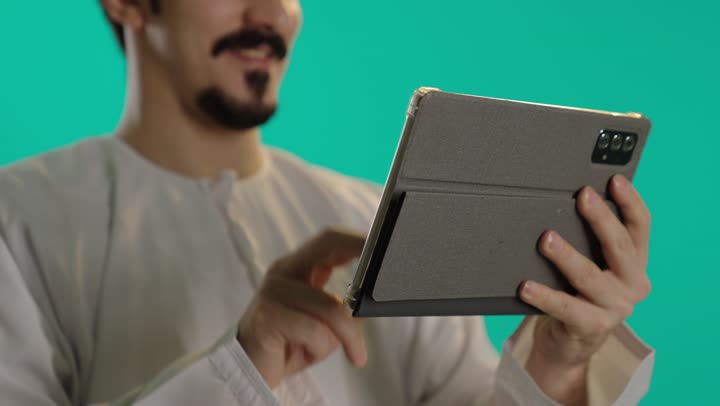 Carrying out the daily required tasks with passion, an Arab Gulf Omani man wearing a white dishdasha and an embroidered kumma holds a tablet easily and lightly, with an enthusiastic and passionate expression on his face.