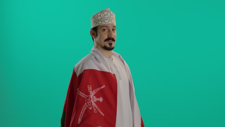 رفع العلم العماني على الاكتاف و نظرة الفخر , رجل عربي عماني خليجي يرتدي الدشداشة البيضاء و الكمة المطرزة يحمل الراية العمانية بثبات و ثقة , تعابير السعادة و الابتسام