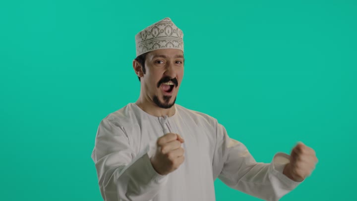 تعبير عن البهجة لتحقيق الفوز , رجل عربي خليجي عماني يرتدي كمة مطرزة و دشداشة بيضاء يحرك قبضة اليدين في الهواء اشارة التهليل و الحماس , اصدار اصوات مرتفعة بالفم اثناء التشجيع